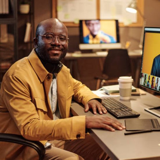 Portrait of African graphic designer in eyeglasses smiling at camera while working at table on computer with new content